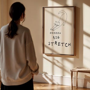 May include: A framed art print featuring a minimalist line drawing of a stretching cat. The artwork includes the words "Ooooooh Big Stretch" in black text. The frame is a warm brown color, and the background is a neutral beige.