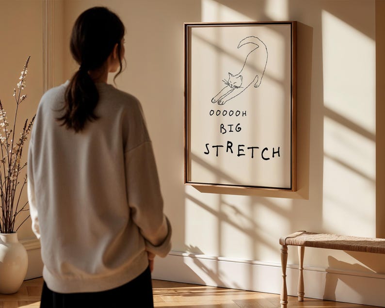 May include: A framed art print featuring a minimalist line drawing of a stretching cat. The artwork includes the words "Ooooooh Big Stretch" in black text. The frame is a warm brown color, and the background is a neutral beige.