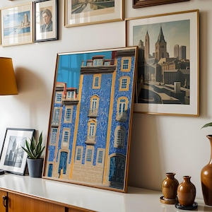 K&ouml;nnte beinhalten: Ein gerahmter Kunstdruck eines blauen Geb&auml;udes mit gelben Akzenten und Balkonen. Das Kunstwerk wird auf einer wei&szlig;en Oberfl&auml;che zusammen mit anderen gerahmten Bildern, einer Lampe und Vasen pr&auml;sentiert. Thema: Wohnkultur.