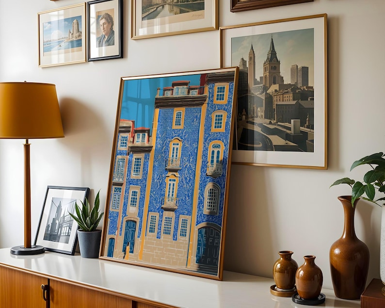 K&ouml;nnte beinhalten: Ein gerahmter Kunstdruck eines blauen Geb&auml;udes mit gelben Akzenten und Balkonen. Das Kunstwerk wird auf einer wei&szlig;en Oberfl&auml;che zusammen mit anderen gerahmten Bildern, einer Lampe und Vasen pr&auml;sentiert. Thema: Wohnkultur.