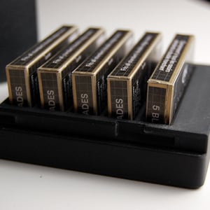 Puede incluir: Una caja de plástico negra con seis cartuchos de película de color dorado. Los cartuchos están etiquetados con "ADES" y "5 BL", y tienen un diseño en blanco y negro.