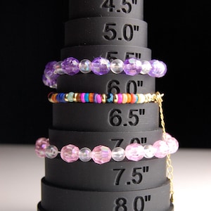 May include: A set of six black silicone bracelet sizing rings with measurements from 4.5 inches to 8 inches.  The rings are stacked on top of each other.  Three bracelets are displayed on the rings: a purple beaded bracelet, a multi-colored beaded bracelet, and a pink beaded bracelet.