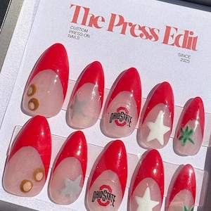 Könnte beinhalten: Ein Set individueller Press-On-Nägel mit rotem und transparentem Design. Einige Nägel zeigen das Ohio State-Logo, weiße Sterne oder braune Kreise. Der Text "The Press Edit" und "Custom Press-On Nails" ist sichtbar.
