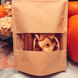May include: A tan paper pouch with a clear rectangular window displaying dried apple slices, cinnamon sticks, and whole cloves. The pouch is sealed at the top and features a flat bottom, containing a mix of dried fruit and spices for aromatic use.