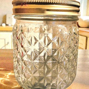 May include: A clear glass jar with a decorative starburst pattern and a gold-coloured metal lid. The jar is designed for preserving food items and has a wide mouth for easy filling and access.