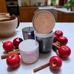 Könnte beinhalten: Duftkerzen in metallischen Behältern, mit einer Kerze mit weißem Deckel im Vordergrund. Rote Äpfel und Zimtstangen sind um die Kerzen angeordnet. Auf dem Kerzenetikett steht "Daybreak Candle Creations".