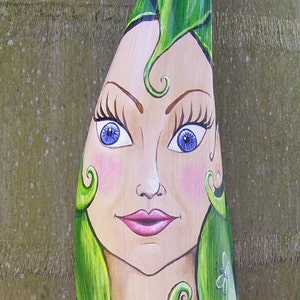 May include: A whimsical wooden carving of a woman with green hair and blue eyes. The carving is painted with details like pink cheeks and a pink mouth. The woman's hair is styled in a curly, green design.
