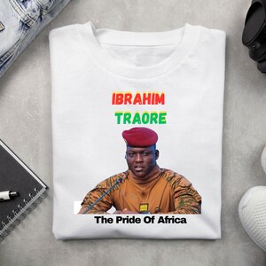 May include: White t-shirt featuring a portrait of Ibrahim Traore with the text "IBRAHIM TRAORE" in red, green, and yellow, and "The Pride Of Africa" below. The shirt is folded on a light gray surface.