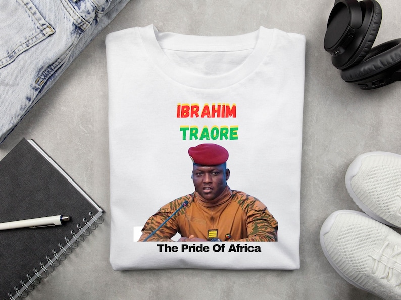 May include: White t-shirt featuring a portrait of Ibrahim Traore with the text "IBRAHIM TRAORE" in red, green, and yellow, and "The Pride Of Africa" below. The shirt is folded on a light gray surface.