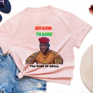 May include: Light pink t-shirt featuring a portrait of Ibrahim Traore with the text "IBRAHIM TRAORE" in red and green, and "The Pride of Africa" below. The shirt is styled with denim shorts, sunglasses, and sandals.