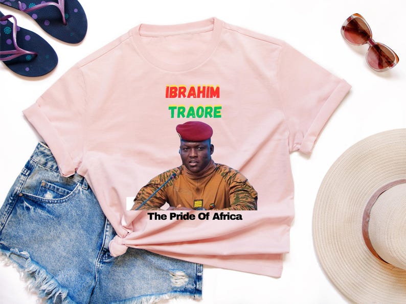 May include: Light pink t-shirt featuring a portrait of Ibrahim Traore with the text "IBRAHIM TRAORE" in red and green, and "The Pride of Africa" below. The shirt is styled with denim shorts, sunglasses, and sandals.
