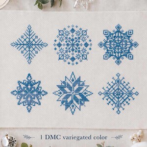 May include: A cross stitch pattern on light fabric featuring six distinct blue snowflake designs arranged in two rows. The text "PDF pattern" appears in a circular badge, and the text "1 DMC variegated color" is printed below the designs.