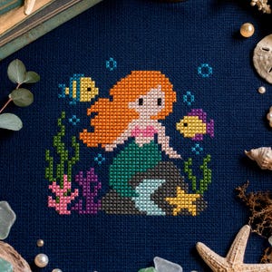 Può includere: Un ricamo a punto croce su tessuto blu scuro raffigurante una sirena con capelli arancioni, coda verde e top rosa, circondata da due pesciolini, alghe verdi, coralli rosa e viola e una stella marina gialla in uno stile pixelato.