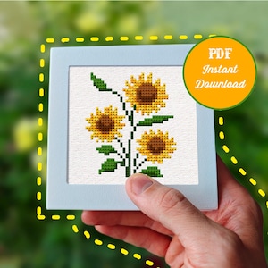 May include: A cross-stitch pattern featuring three yellow sunflowers with brown centers and green leaves on a white fabric background, presented in a square white frame with a circular orange badge that reads PDF Instant Download.