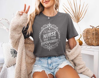 May include: Dark gray t-shirt with the text "I'M A NURSE WHAT'S YOUR SUPERPOWER?" in a white decorative frame. The shirt is worn with denim shorts and a beige cardigan. A peace sign is made with the fingers.