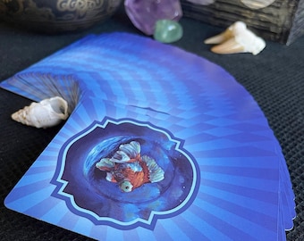Könnte beinhalten: Ein Tarotkartenspiel mit einem blau-weißen Design mit einem Fisch in einem Wirbelmuster. Die Karten sind aufgefächert, mit einer Muschel und Kristallen im Hintergrund. Eine bronzene Klangschale ist ebenfalls sichtbar.