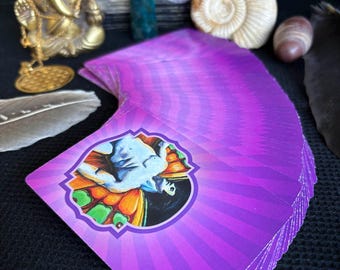 Könnte beinhalten: Ein Tarotkartendeck mit einem lila und weißen Design, das eine Illustration einer Figur in einem kreisförmigen Rahmen zeigt. Die Karten sind aufgefächert und zeigen die detaillierten Kunstwerke. Weitere Artikel sind eine goldene Statue, eine Muschel und eine Feder.