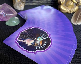 Könnte beinhalten: Ein Tarotkartenspiel mit einem lila und weißen Design. Die Karten zeigen eine Illustration eines Frauengesichts mit einer Blume und anderen dekorativen Elementen. Die Karten sind auf einer schwarzen Oberfläche aufgefächert, mit Kristallen und einer goldenen Statue im Hintergrund.
