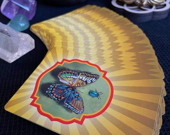 Könnte beinhalten: Ein Tarotkartenspiel mit einer Illustration eines Schmetterlings und eines Käfers. Die Karten haben auf der Rückseite ein gelbes und goldenes Sonnenstrahlendesign. Die Illustration zeigt einen Schmetterling mit blauen, orangefarbenen und grünen Flügeln.