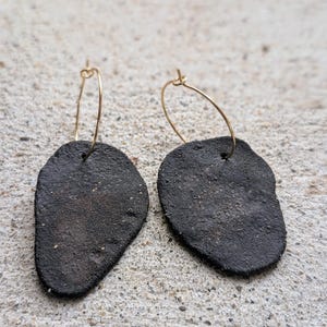 Puede incluir: Un par de pendientes de forma orgánica gris oscuro con cierres de aro dorados. Los pendientes tienen una superficie texturizada y una estética natural y terrosa. Los pendientes se muestran sobre una superficie gris claro.