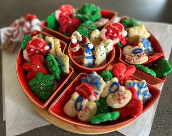 May include: A festive assortment of Christmas cookies in a red divided serving dish. The cookies are decorated with red, green, white, and blue icing, featuring shapes like snowmen, trees, and hearts. A wooden tray holds the dish.