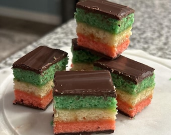 May include: A stack of square Italian rainbow cookies featuring three distinct layers of almond sponge cake in green, white, and orange, topped with a smooth dark chocolate glaze. Each small rectangular treat is layered with fruit preserves.