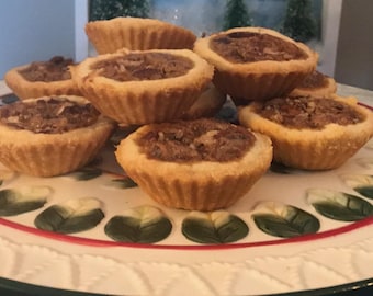 May include: A stack of mini pecan pies on a decorative plate. The pies have a golden-brown crust and a filling of pecans. The plate has a white background with green and white leaf designs and a red border.