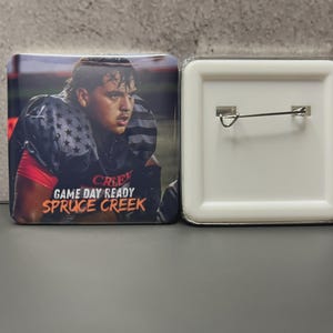 May include: Square button featuring a football player in a dark jersey with stars and stripes. The text "GAME DAY READY" and "SPRUCE CREEK" are in orange. The button has a white back with a pin.