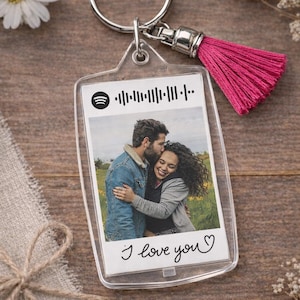 May include: A clear acrylic keychain with a photo of a couple embracing, a Spotify code, and the handwritten text "I love you". The keychain has a silver ring and a pink tassel. The background is a wooden surface.