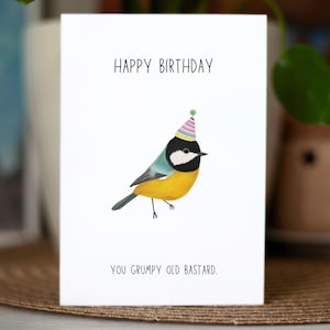 May include: A white greeting card with the text "HAPPY BIRTHDAY" at the top and "YOU GRUMPY OLD BASTARD." at the bottom. The card features a colorful illustration of a bird wearing a party hat.