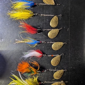 May include: Eight gold fishing lures with different coloured feathers and beads. The lures are arranged vertically on a dark grey surface.