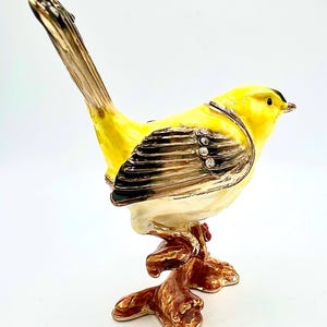 May include: A yellow bird figurine with black accents and rhinestones perched on a brown branch. The bird has a long tail and a small beak. The figurine is made of metal and has a shiny finish.