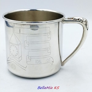 Può includere: Una tazza color argento lucido con l'incisione di una torre dell'orologio e una pergamena vuota con il testo lbs. oz. La tazza presenta un corpo cilindrico liscio e un manico curvo con dettagli decorativi nel punto di attacco superiore.