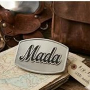 May include: A rectangular silver-toned metal belt buckle featuring the word Mada in raised black script lettering. The buckle is positioned on top of a vintage-style map, with a brown leather saddle and a leather bag visible in the background.
