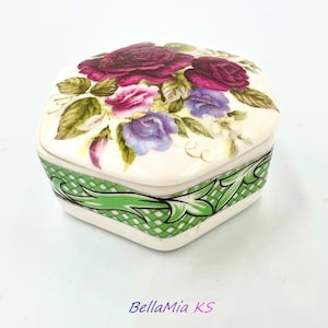 May include: A small, square trinket box made of white ceramic with a floral design on the lid. The lid features a bouquet of pink, purple, and red roses with green leaves. The box has a green and white checkered border.