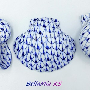 Könnte beinhalten: Drei weiße Keramik-Muschelfiguren mit blauem Fischernetzmuster. Die Muscheln haben unterschiedliche Formen, darunter eine Spirale und eine Jakobsmuschel. Der Text "BellaMia KS" befindet sich unten.