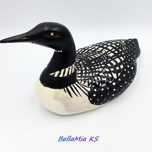 May include: A hand-painted wooden loon figurine. The bird has a black head and neck with white stripes, a white belly, and a black and white patterned back. The text "BellaMia KS" is at the bottom.