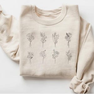 May include: A beige sweatshirt with a graphic design of nine different floral bouquets in black line art.
