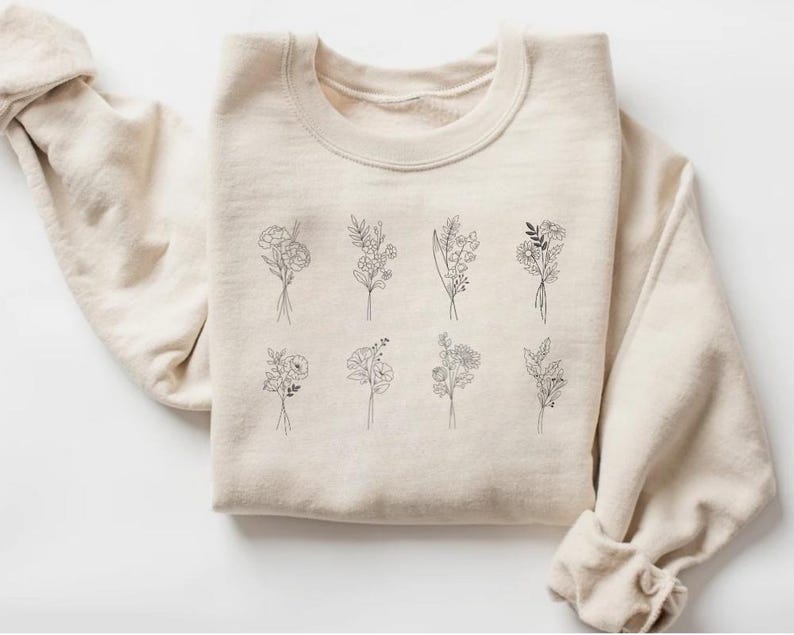 May include: A beige sweatshirt with a graphic design of nine different floral bouquets in black line art.