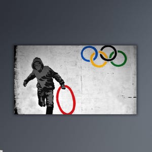 May include: A framed print featuring a stencil-style image of a person running with a red ring, set against a distressed white background. The Olympic rings are in the upper right corner. The artwork is mounted on a gray wall.
