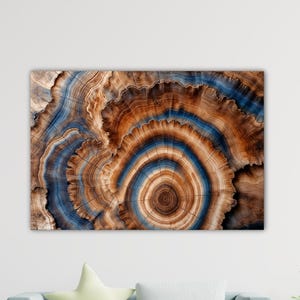Op de afbeelding: Abstracte kunstprint met een close-up van een dwarsdoorsnede van een boomstam. Het kunstwerk toont concentrische ringen in tinten bruin, blauw en beige, waardoor een natuurlijk, organisch patroon ontstaat. De print is aan een muur bevestigd.