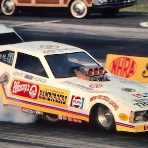 May include: A white dragster with yellow and red accents, featuring a Wendy's logo and the text "You've Had The Taste" on the side. The car is in motion, leaving a trail of smoke behind it.