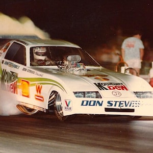 May include: A white drag racing car with a Mountain Dew sponsorship. The car is in motion and leaving a trail of smoke. The car has the text "DON STEVENS" on the front.