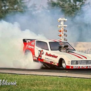 May include: A white and red race car with the word "Budweiser" on the side, is doing a burnout, leaving a large cloud of smoke behind it. The car has the number "12" on the side and a cartoon character on the door.