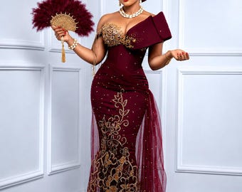 May include: A floor-length burgundy gown featuring an off-the-shoulder neckline, gold sequin embroidery, and a sheer tulle overlay. The ensemble includes a matching burgundy headpiece, a beaded necklace, and a handheld burgundy feather fan.
