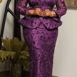 May include: A deep purple two-piece ensemble featuring a long-sleeved peplum top and a matching floor-length skirt. The garment is crafted from intricate floral lace fabric with a scalloped hemline on the sleeves and a fitted silhouette.