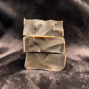 Pode incluir: Uma pilha de três barras de sabão retangulares com uma cor castanho-taupe sólida, suave e discreta. Cada barra apresenta variações tonais subtis e onduladas na sua superfície, criando um efeito marmoreado. As bordas são retas e limpas.