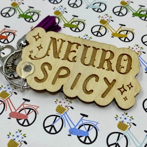 Puede incluir: Llavero de madera con el texto "Neuro Spicy" en una fuente decorativa. El llavero tiene forma de nube con un dise&ntilde;o brillante. El fondo es un patr&oacute;n blanco de bicicletas con flores.