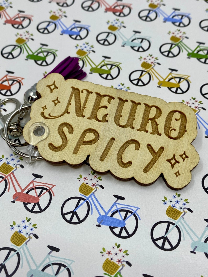 Puede incluir: Llavero de madera con el texto "Neuro Spicy" en una fuente decorativa. El llavero tiene forma de nube con un dise&ntilde;o brillante. El fondo es un patr&oacute;n blanco de bicicletas con flores.