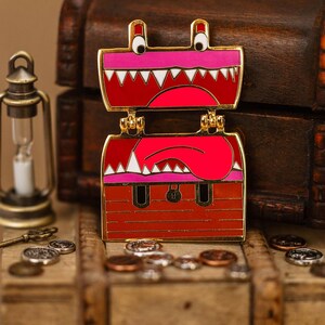 May include: A red and pink enamel pin in the shape of an open treasure chest with a cartoon monster face. The pin has gold-colored accents and is set against a wooden background with coins and a lantern.